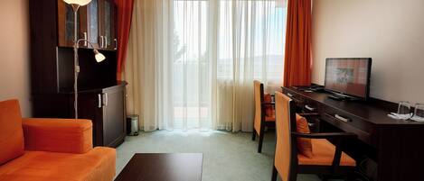 Una habitación de hotel con sofá, escritorio, silla, televisión y ventana con cortinas.