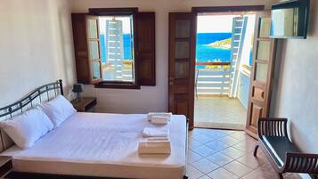 A bedroom with a bed, a desk, a chair, a TV, and a view of the sea through an open door.