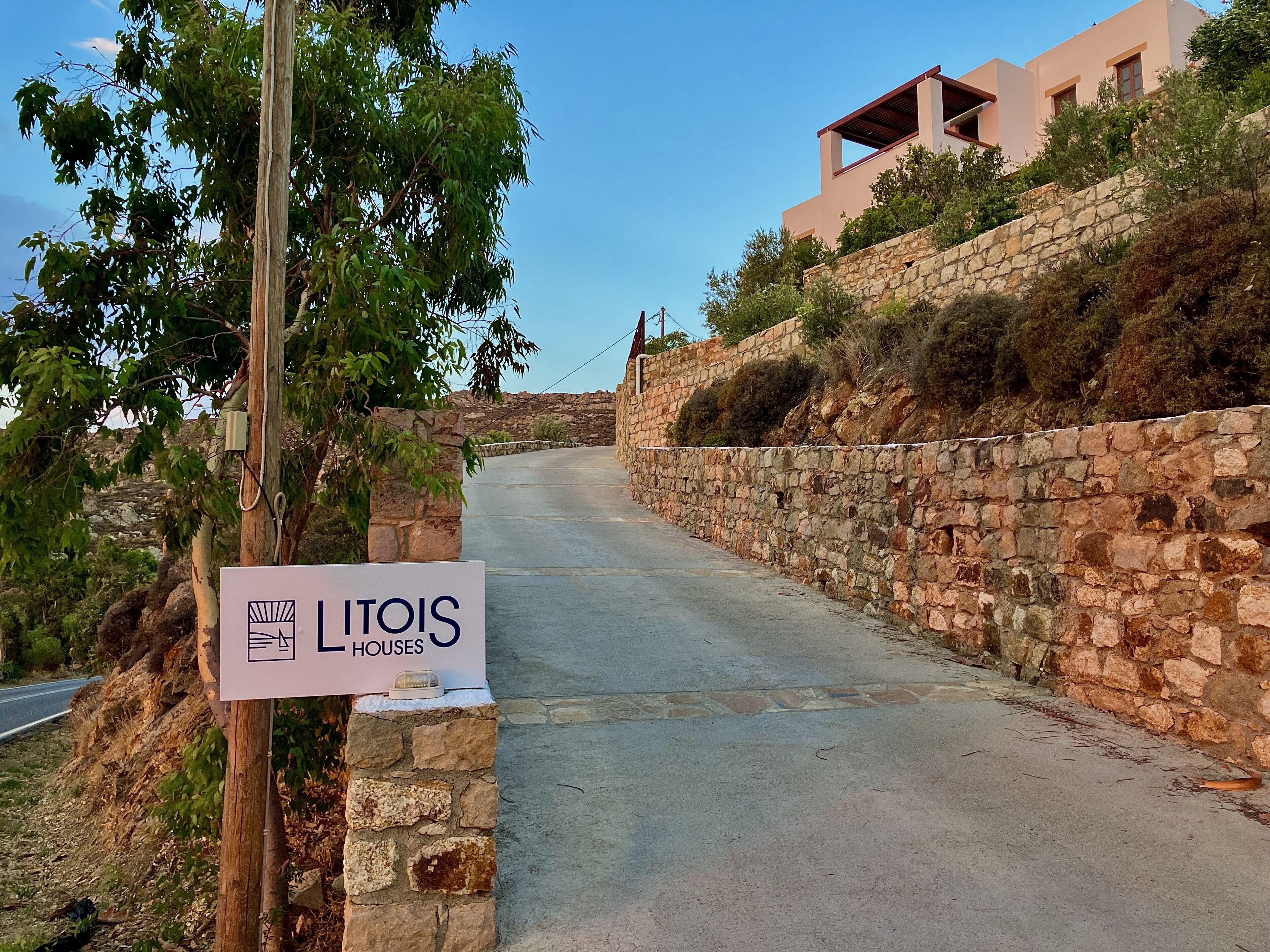 A narrow road flanked by stone walls and greenery, leading to a modern building with a sign reading "Litois Houses".