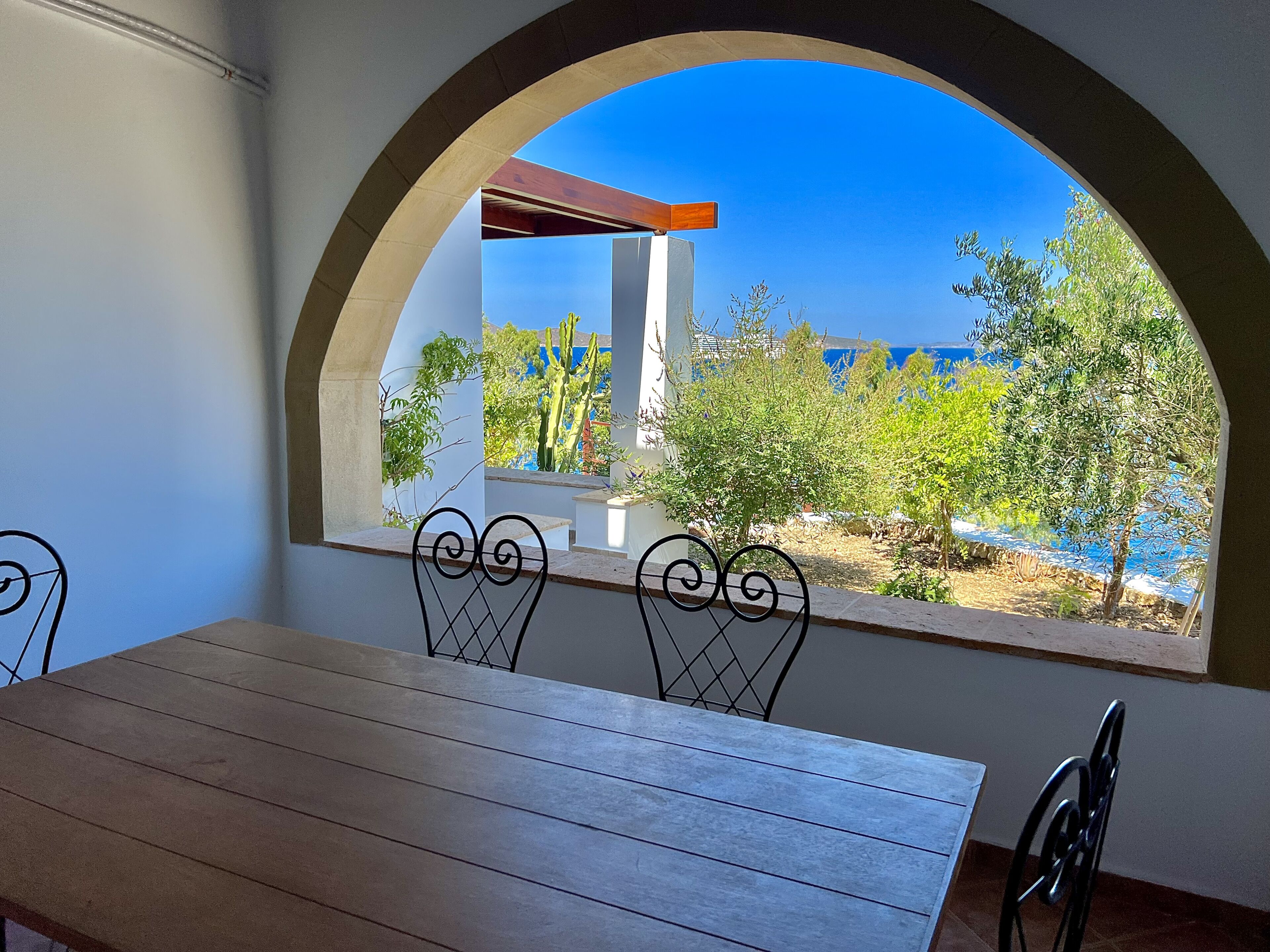 Ein Essbereich mit Blick auf das Meer durch ein Rundbogenfenster, ausgestattet mit einem Holztisch und Metallstühlen.