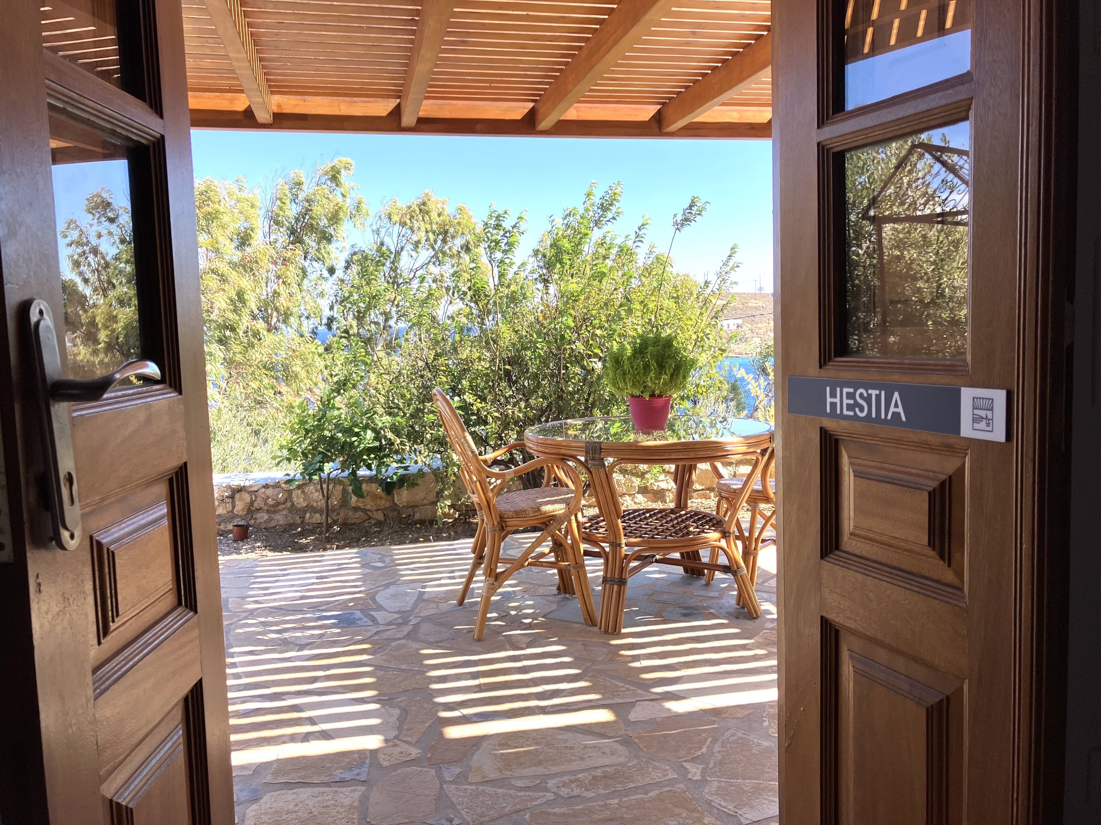 An open wooden door leading to a patio with a stone floor, wicker furniture, and a potted plant.