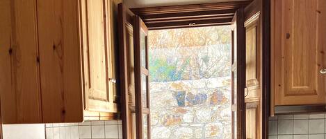 A kitchen with wooden cabinets, a window with a view of a stone wall, a sink, and an oven.