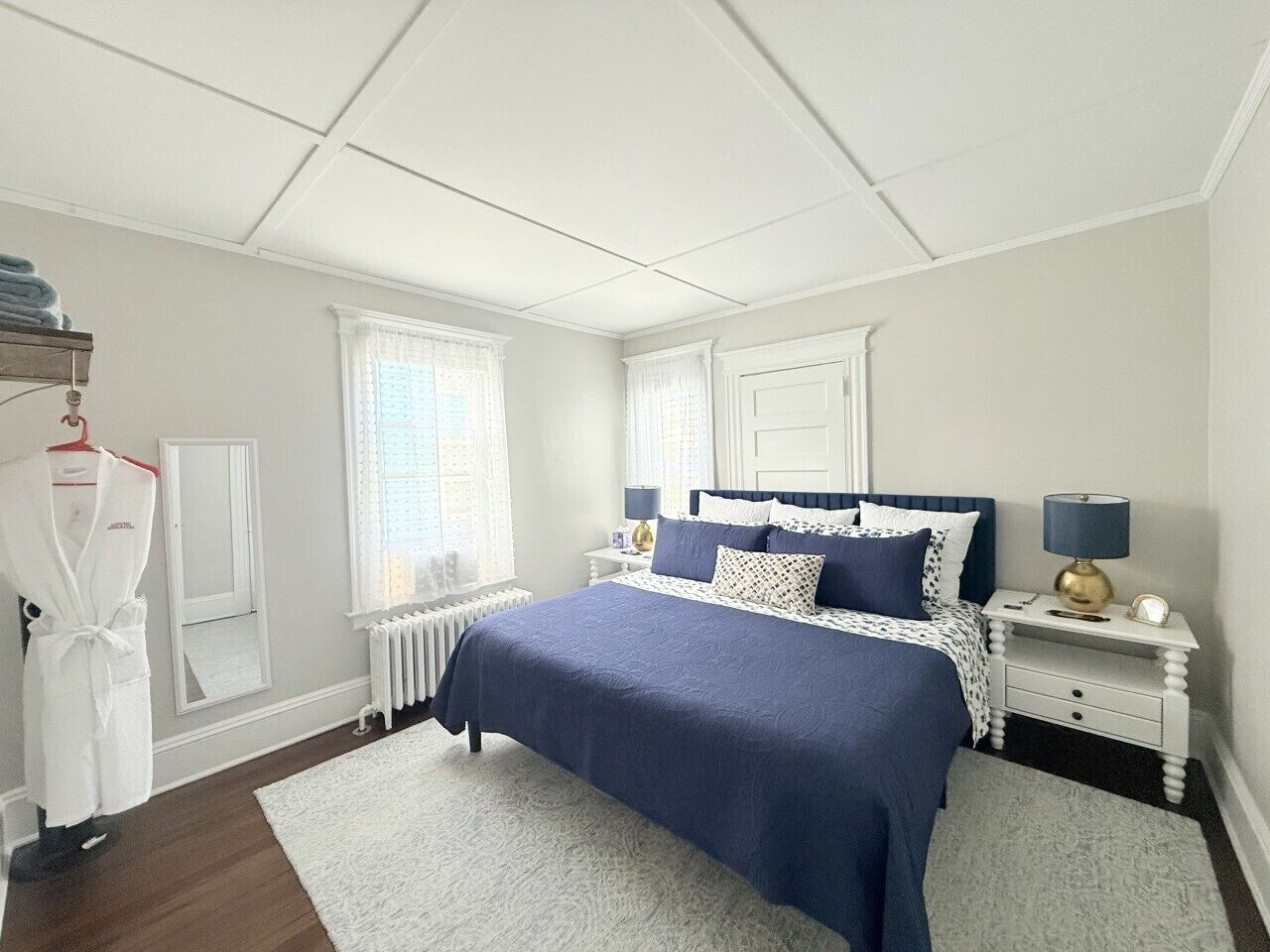 Een slaapkamer met een bed, nachtkastjes, een radiator, een raam met gordijnen en een witte badjas hangend aan een rek.
