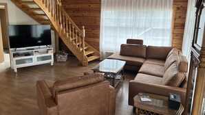 A spacious living room with a wooden staircase, a large flat-screen TV, a brown leather sofa, and a glass coffee table.