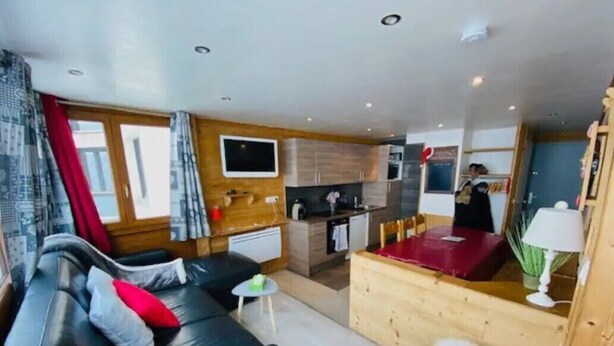 A modern living room with a black leather sofa, a small round table, and a wooden coffee table. There is a kitchen area with wooden cabinets and a red countertop. - Apart. cosy (47m²) - big family room - 3 bedrooms - directly on the slopes (Les Belleville)