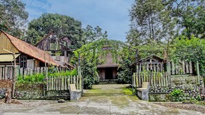 A traditional wooden house with a tiled roof, surrounded by lush greenery and a stone wall. - Hotel O Omah Baturan Syariah (Colomadu)
