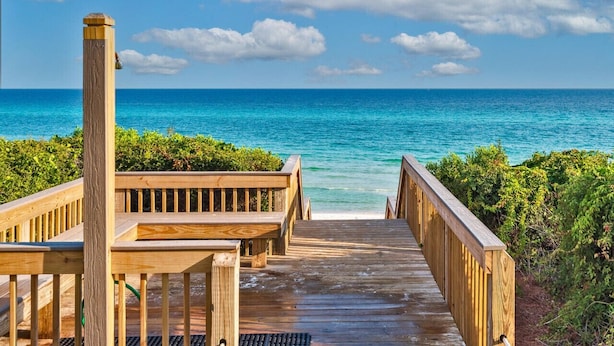 Une terrasse en bois qui mène à une plage où l’eau est d’un bleu limpide et où le ciel est partiellement nuageux.