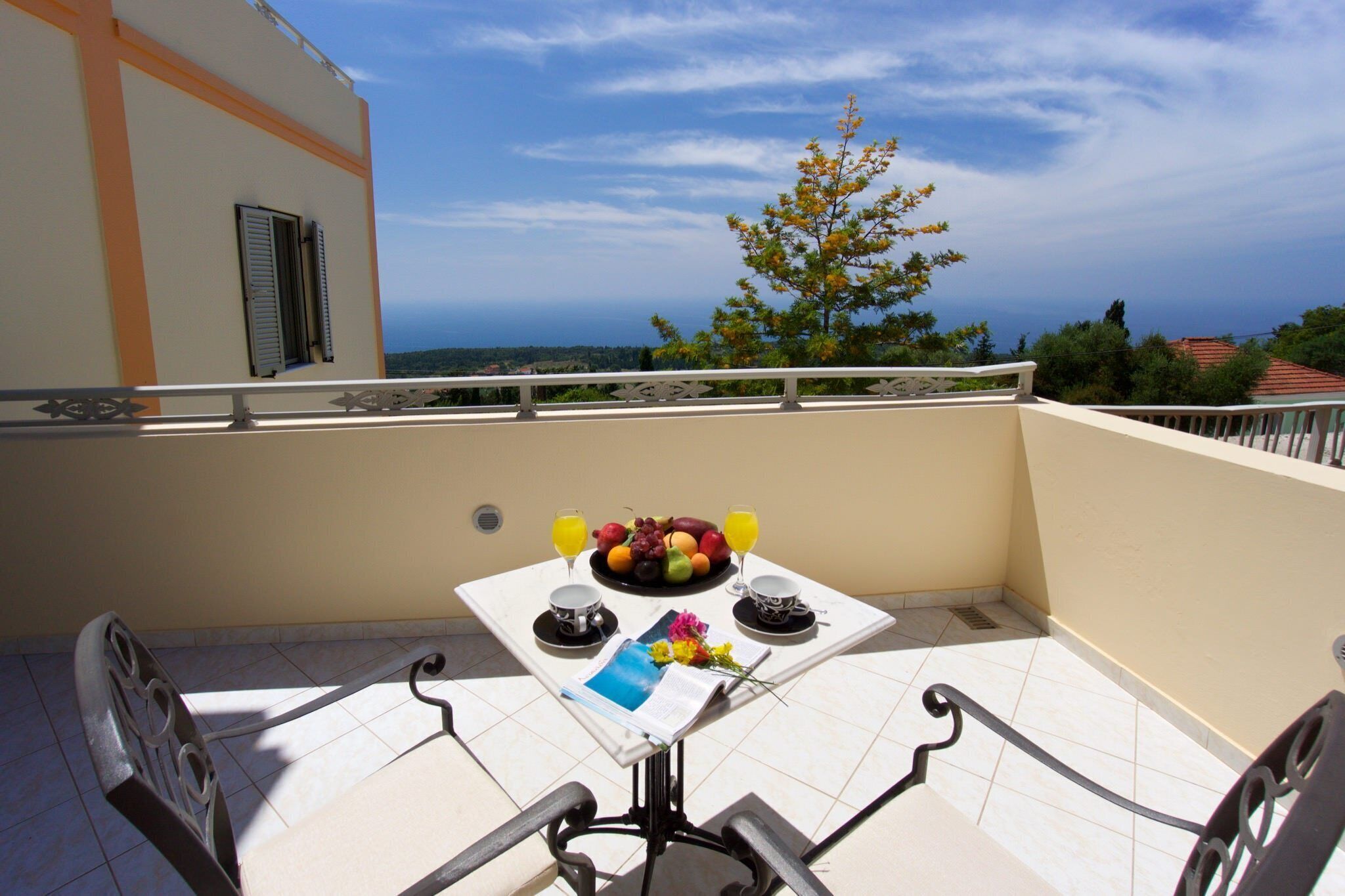 A balcony with a table set for breakfast, including fruits and drinks, overlooking a scenic view.