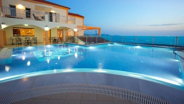 Una piscina de borde curvo, una zona de estar cubierta con sillas y un edificio con balcones que dan a la piscina.