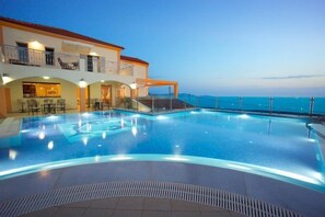 Una piscina de borde curvo, una zona de estar cubierta con sillas y un edificio con balcones que dan a la piscina.