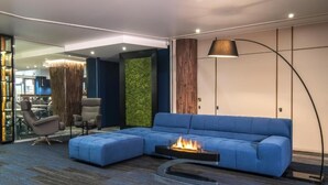 Un salon moderne avec un canapé d’angle bleu, une table basse en verre dotée d’un foyer intégré, un grand lampadaire et une cloison en verre donnant sur une autre pièce.