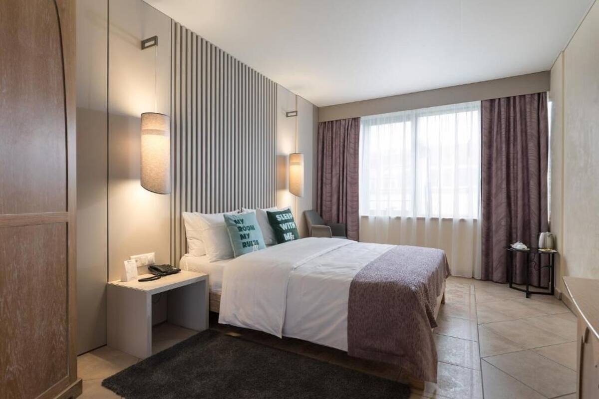A modern hotel room with a neatly made bed, a bedside table with a lamp, a nightstand, and a large window with curtains.