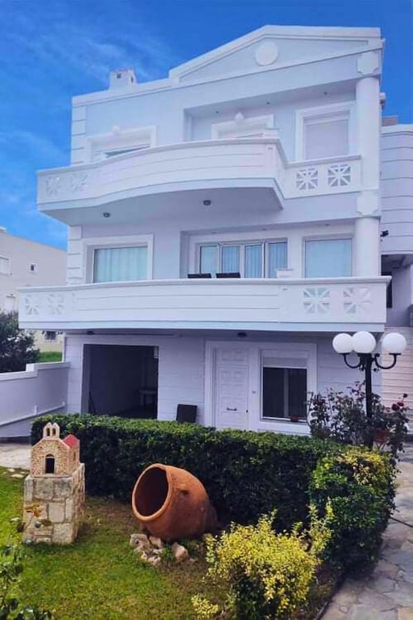 A two-story building with a flat roof, a balcony on the second floor, and a small garden area with a stone structure and a large pot.