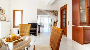 Uma sala ampla com uma mesa de jantar de vidro, cadeiras de vime, um armário de madeira e uma escada que dá para outro cômodo.