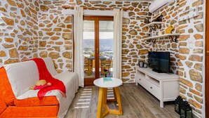 A cozy room with stone walls, a wooden ceiling, a white sofa with a red blanket, a small wooden table, and a TV on a white cabinet. - Olivia's Countryside House (Paros)