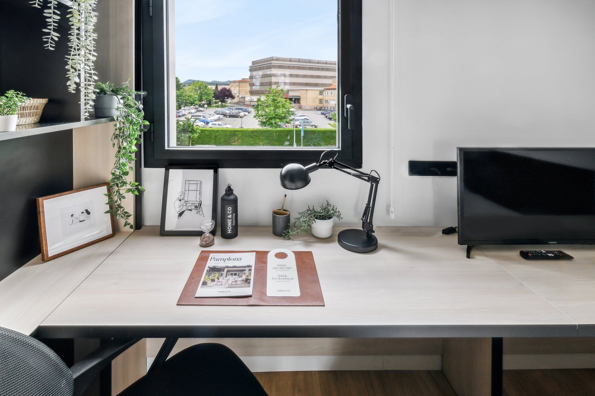 Un bureau moderne, équipé d’un ordinateur et d’une lampe, donnant sur un parking et des immeubles.