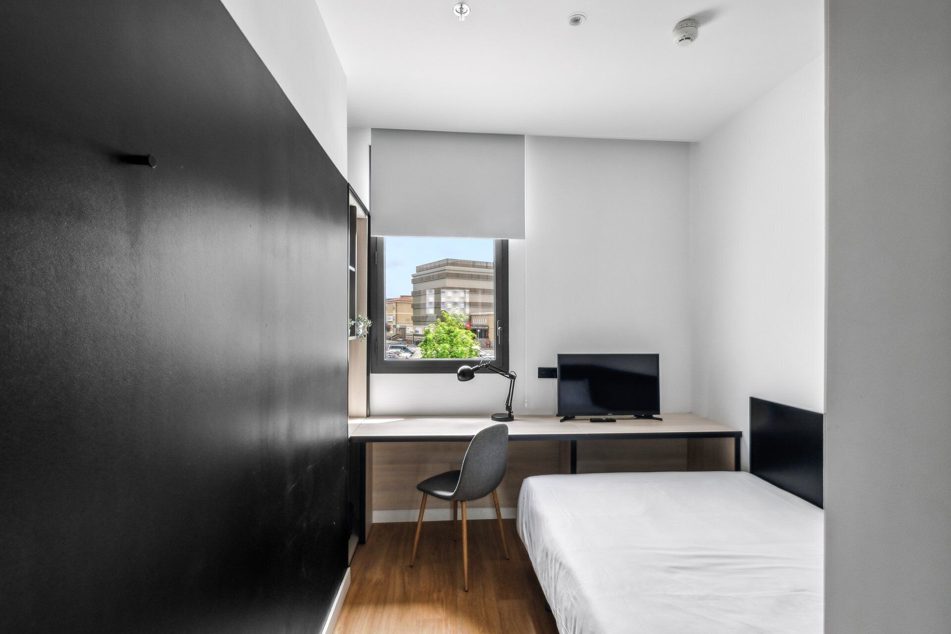 Un dormitorio moderno con cama, escritorio, silla y ventana con vistas a la ciudad.