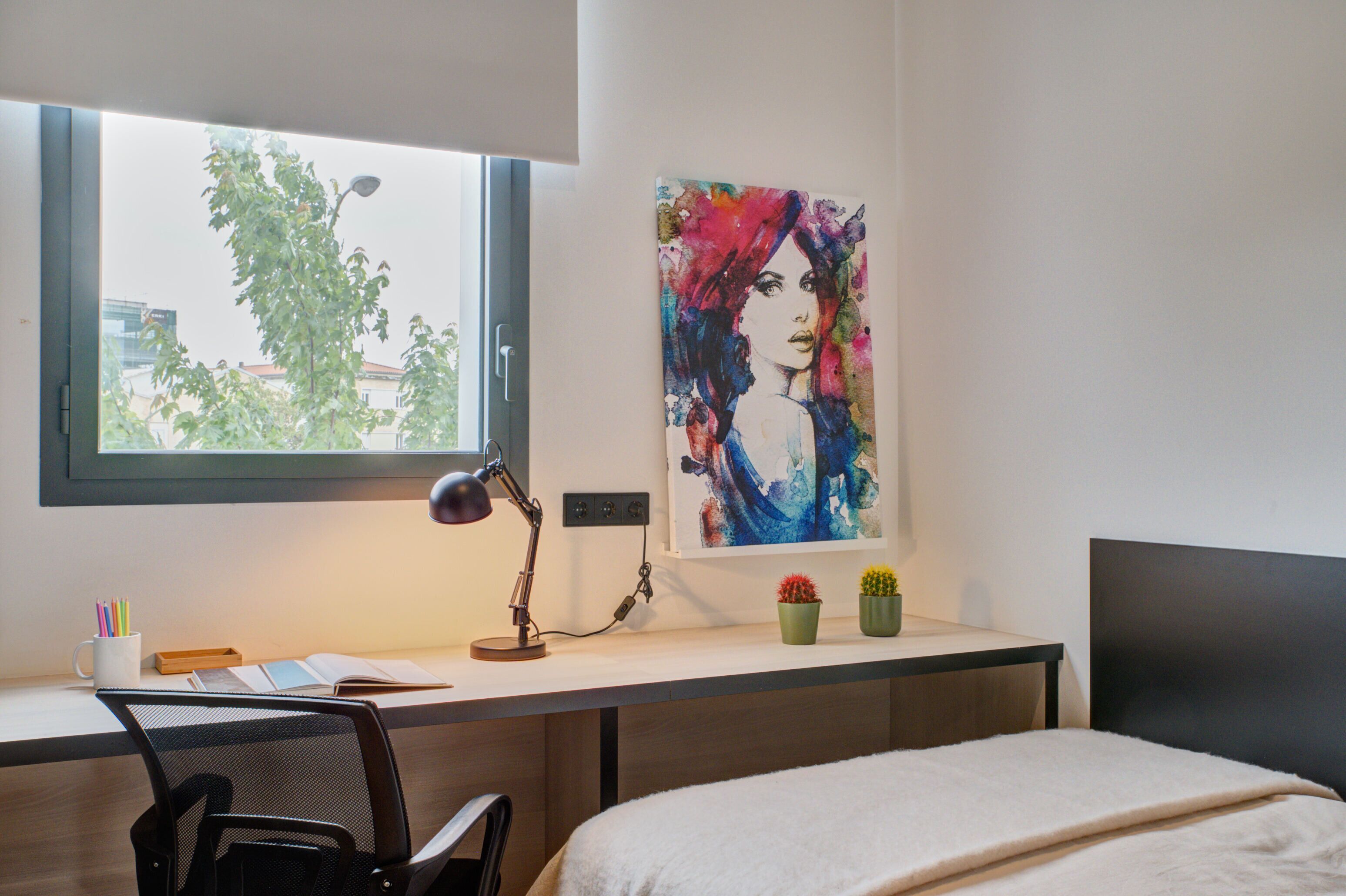 A modern bedroom with a desk, a bed, a window with a view of buildings, and a colorful abstract painting on the wall.