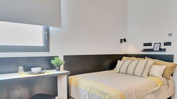 A modern bedroom with a bed, bedside table, and a small desk area with a plant and a glass.