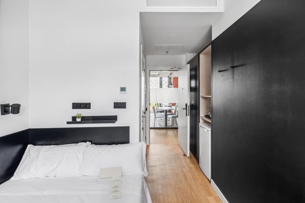Ein modernes Hotelzimmer mit Bett, Nachttisch, einer kleinen Pflanze und Blick in den Küchenbereich.