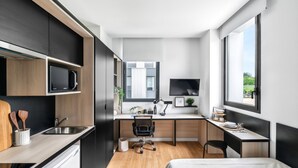 Cozinha compacta e moderna com eletrodomésticos embutidos, área de jantar com escrivaninha e cadeira, e uma janela grande que oferece iluminação natural.