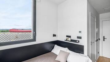 Une chambre d’hôtel moderne avec une grande fenêtre, un lit avec une literie blanche et beige, une tête de lit noire et une petite étagère ornée d’une plante et d’une horloge.