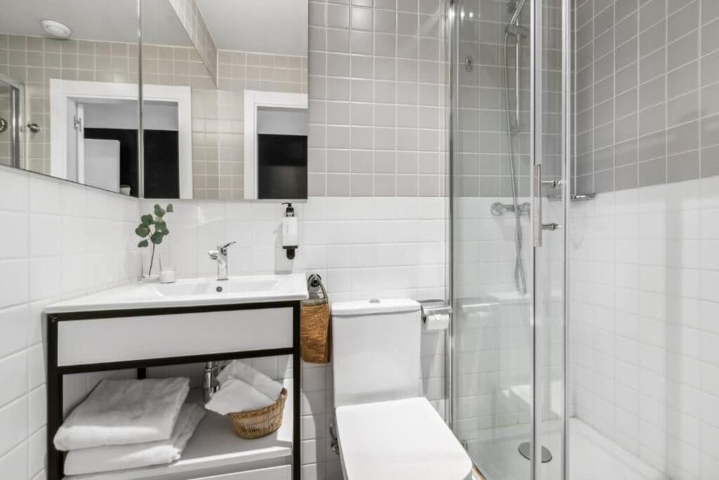 Een moderne badkamer met een douchecabine van glas, witte tegels, een wastafel met spiegel en een toilet.