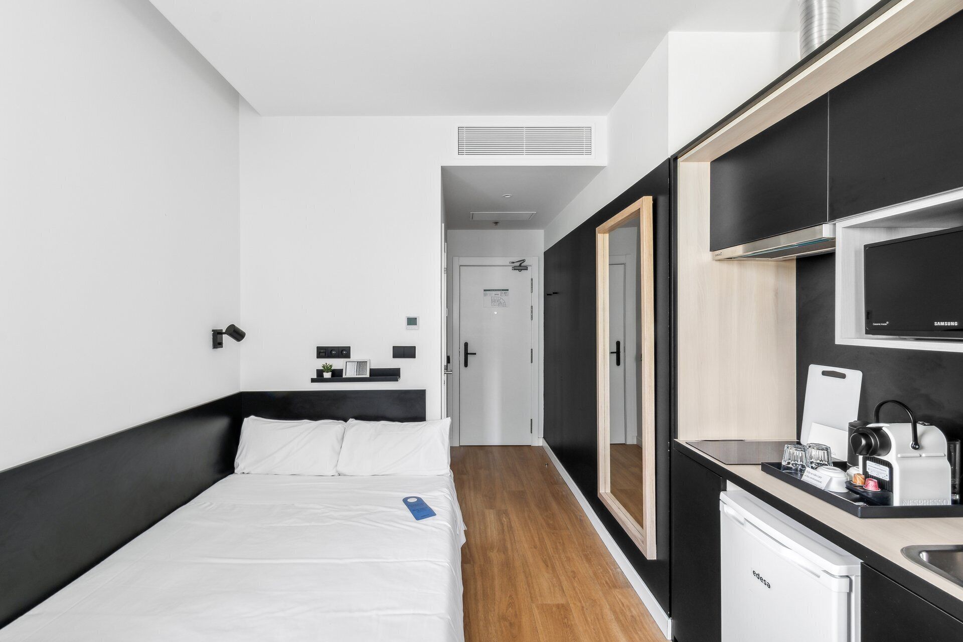 Ein modernes Hotelzimmer mit Bett, Nachttisch und einer Küchenzeile, inklusive Mikrowelle, Kaffeemaschine und kleinem Kühlschrank.