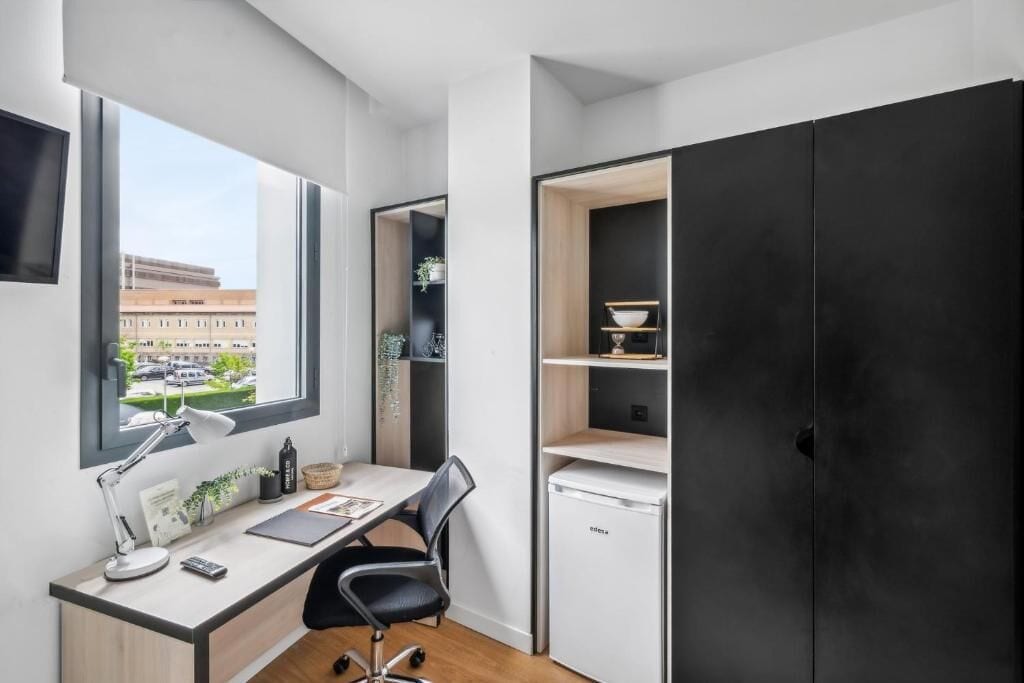 A modern office space with a desk, chair, and a small white refrigerator. There is a window providing a view of buildings outside.