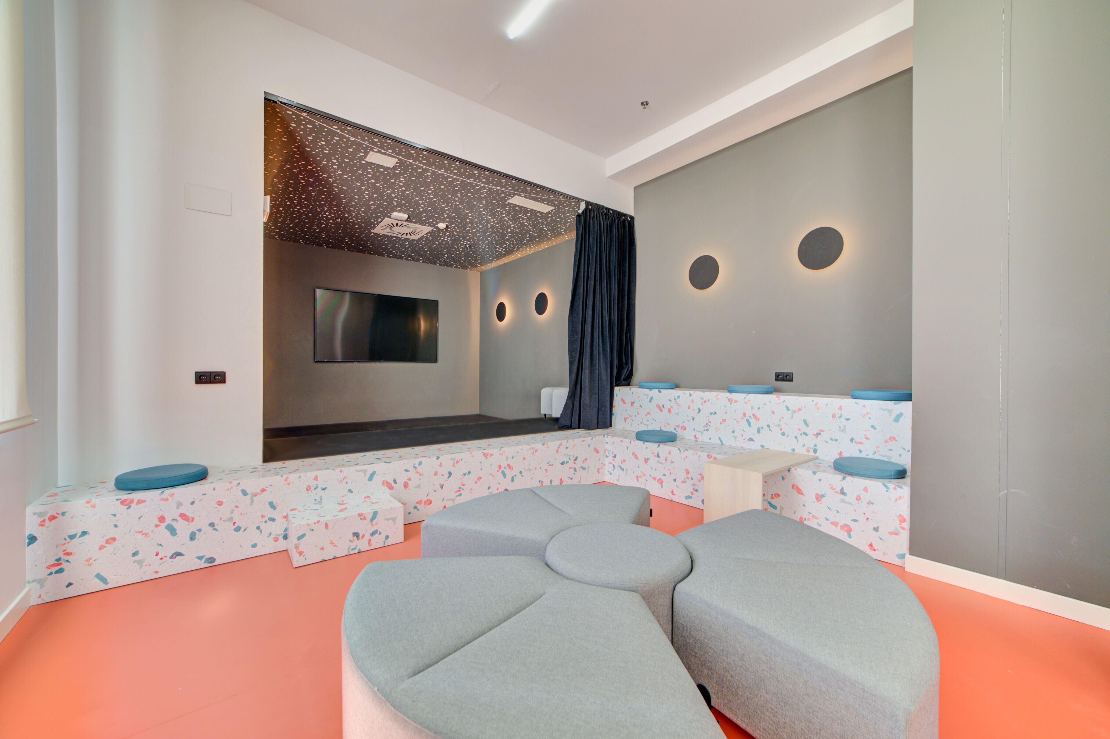 A modern, well-lit room with a seating area featuring a large, curved grey couch and a flat-screen TV mounted on the wall.