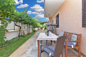 Um pátio externo com mesa e cadeiras, uma pérgola coberta por trepadeiras, e vista para uma rua ao fundo.