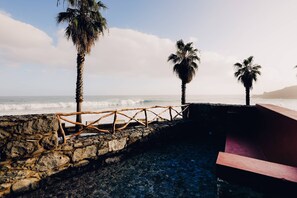 A stone wall with a wooden fence, palm trees, and a view of the ocean. - Casa do Mar - Casas Calhau Miúdo (Seixal)