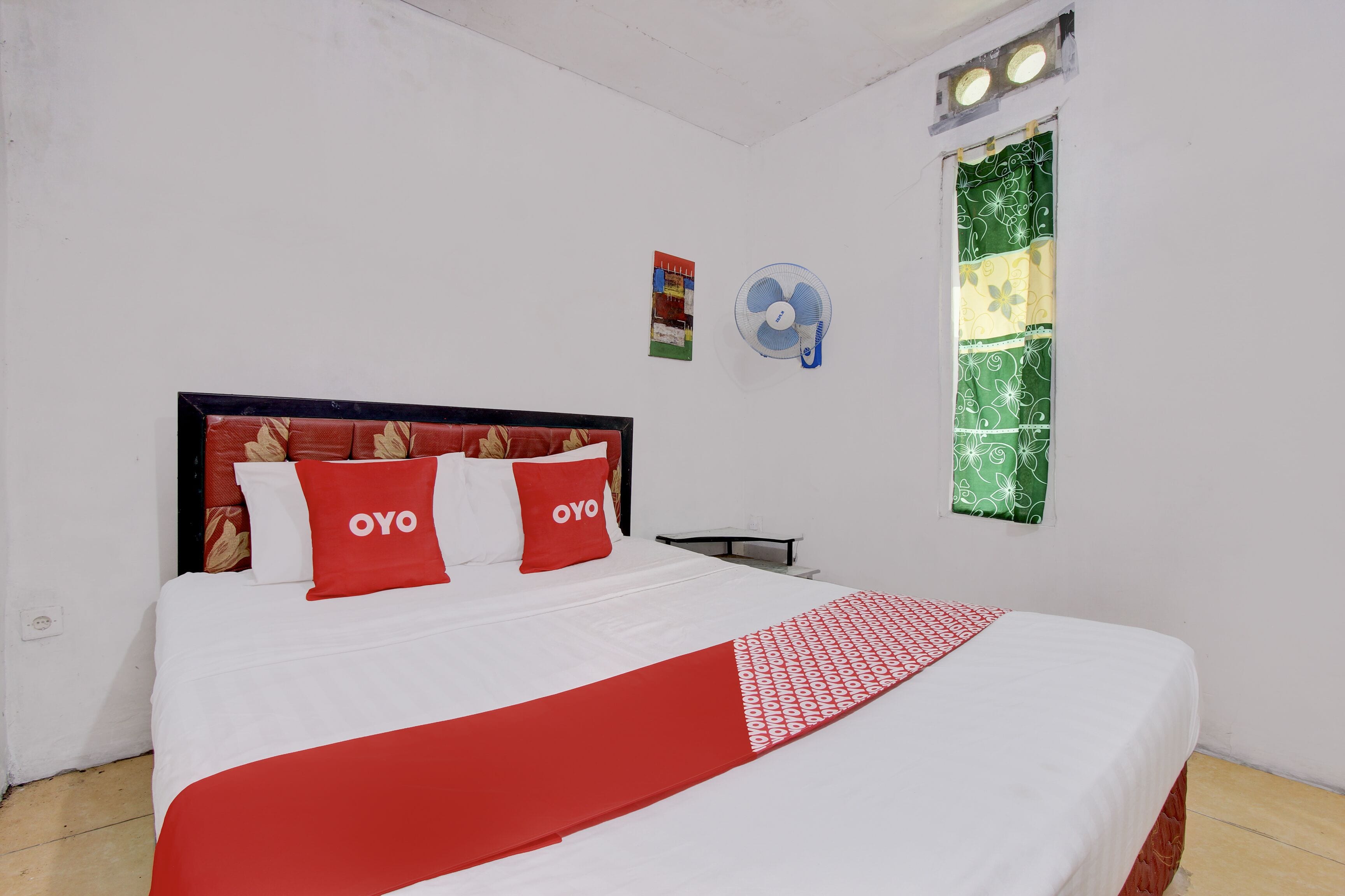 A neatly made bed with a red and white bedspread, two pillows with the word "OYO" on them, a wall-mounted fan, and a window with a green patterned curtain.