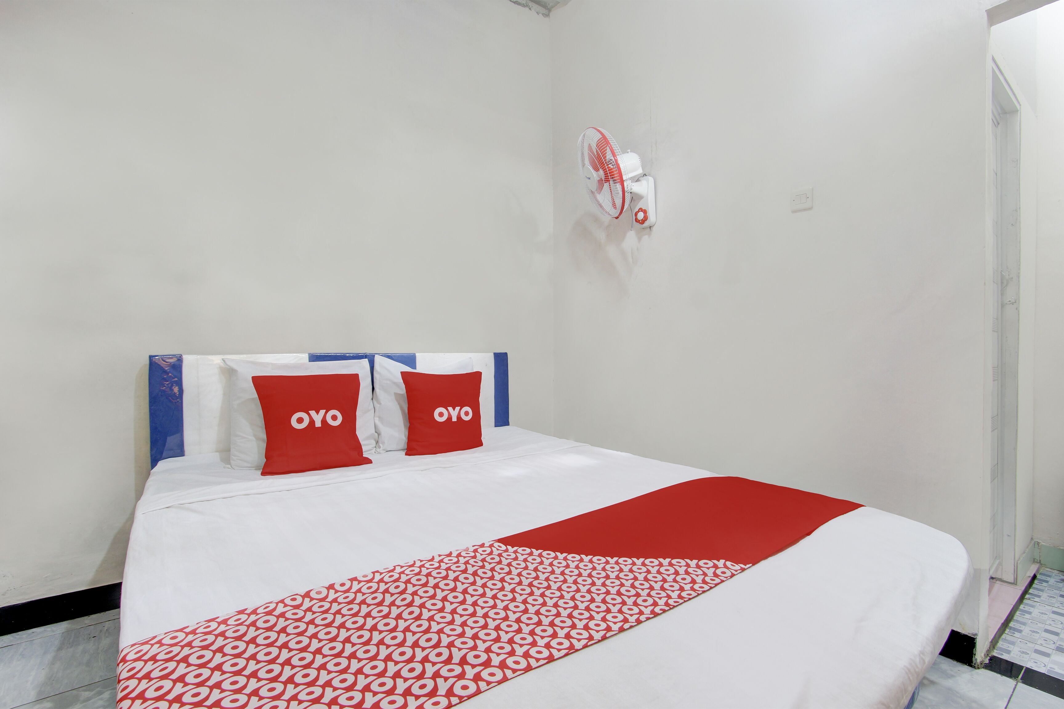 A hotel room with a bed, two red pillows with "OYO" text, a white and red patterned bedspread, and a wall-mounted fan.