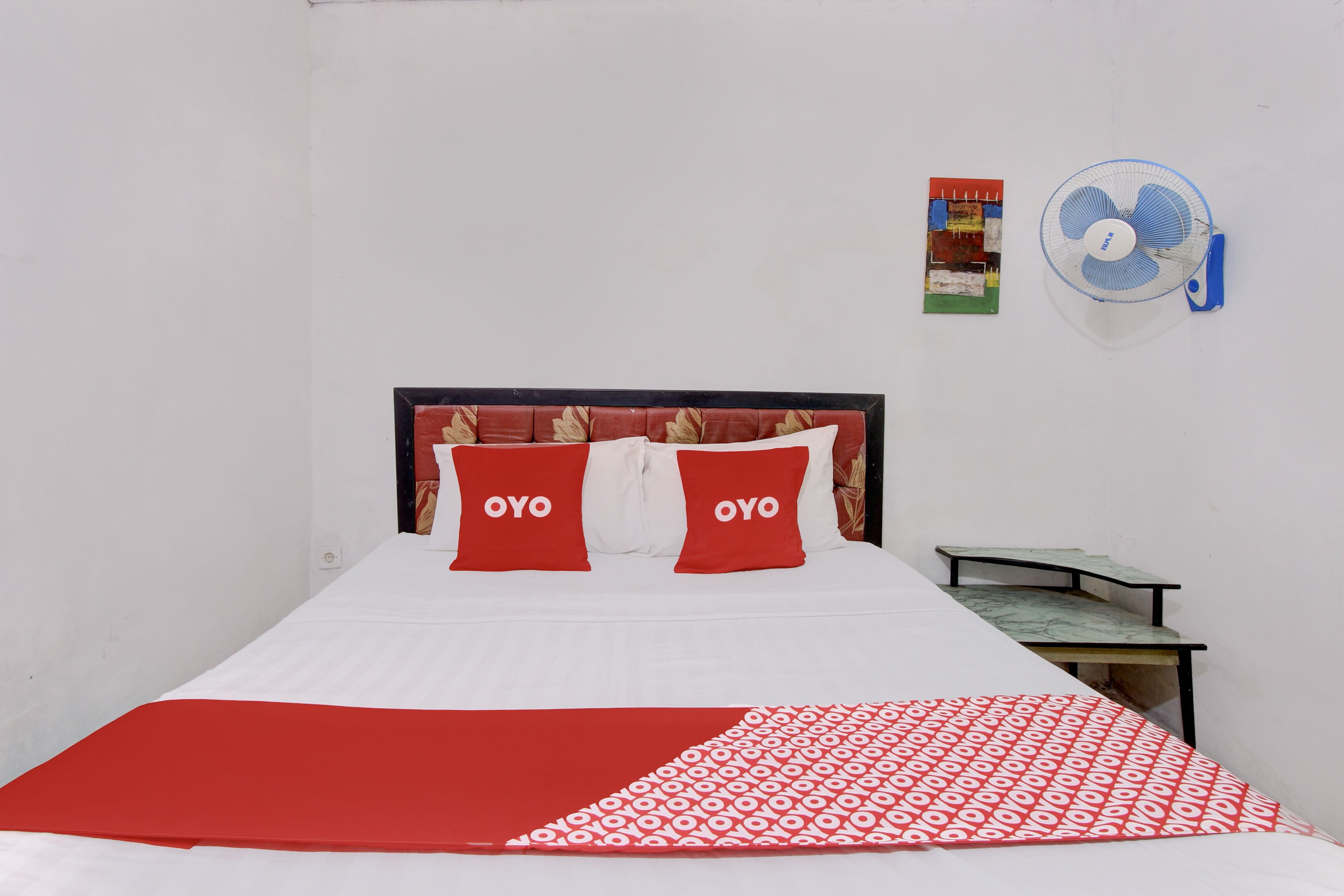 A bed with two pillows labeled "OYO," a small table, a wall-mounted fan, and two posters on the wall.