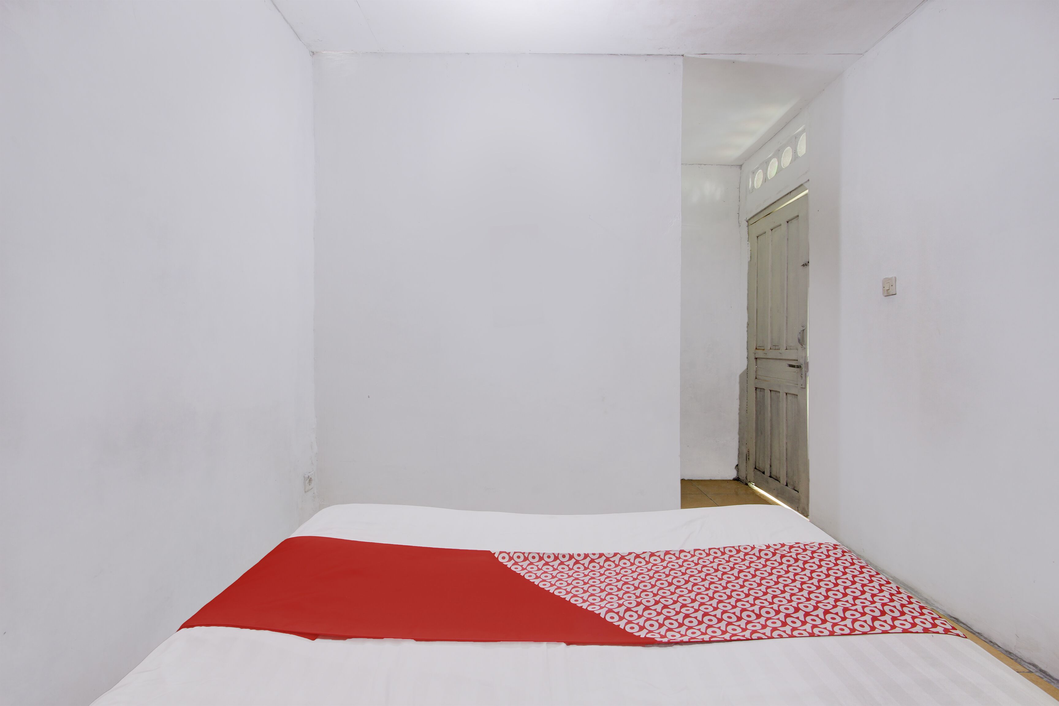 Habitación estrecha con una cama al fondo, paredes blancas y una puerta que da a otra habitación.