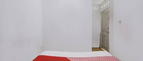 Habitación estrecha con una cama al fondo, paredes blancas y una puerta que da a otra habitación.