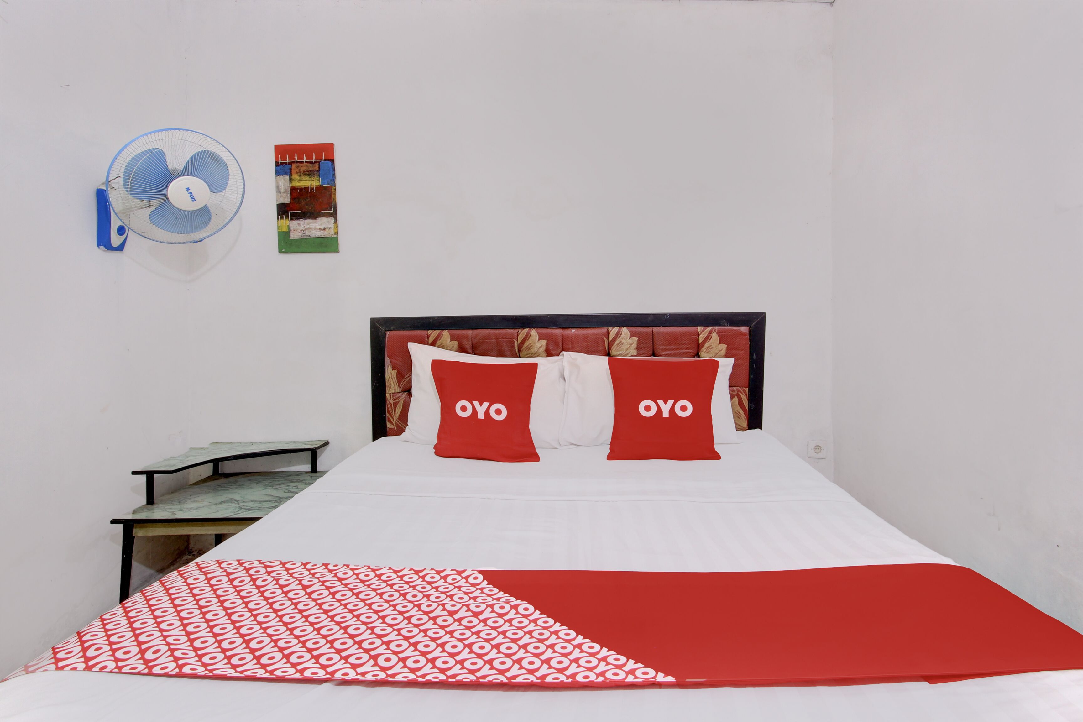 Una cama individual con dos almohadas rojas, una colcha blanca y roja, una mesita, un ventilador de pared y un cuadro enmarcado.