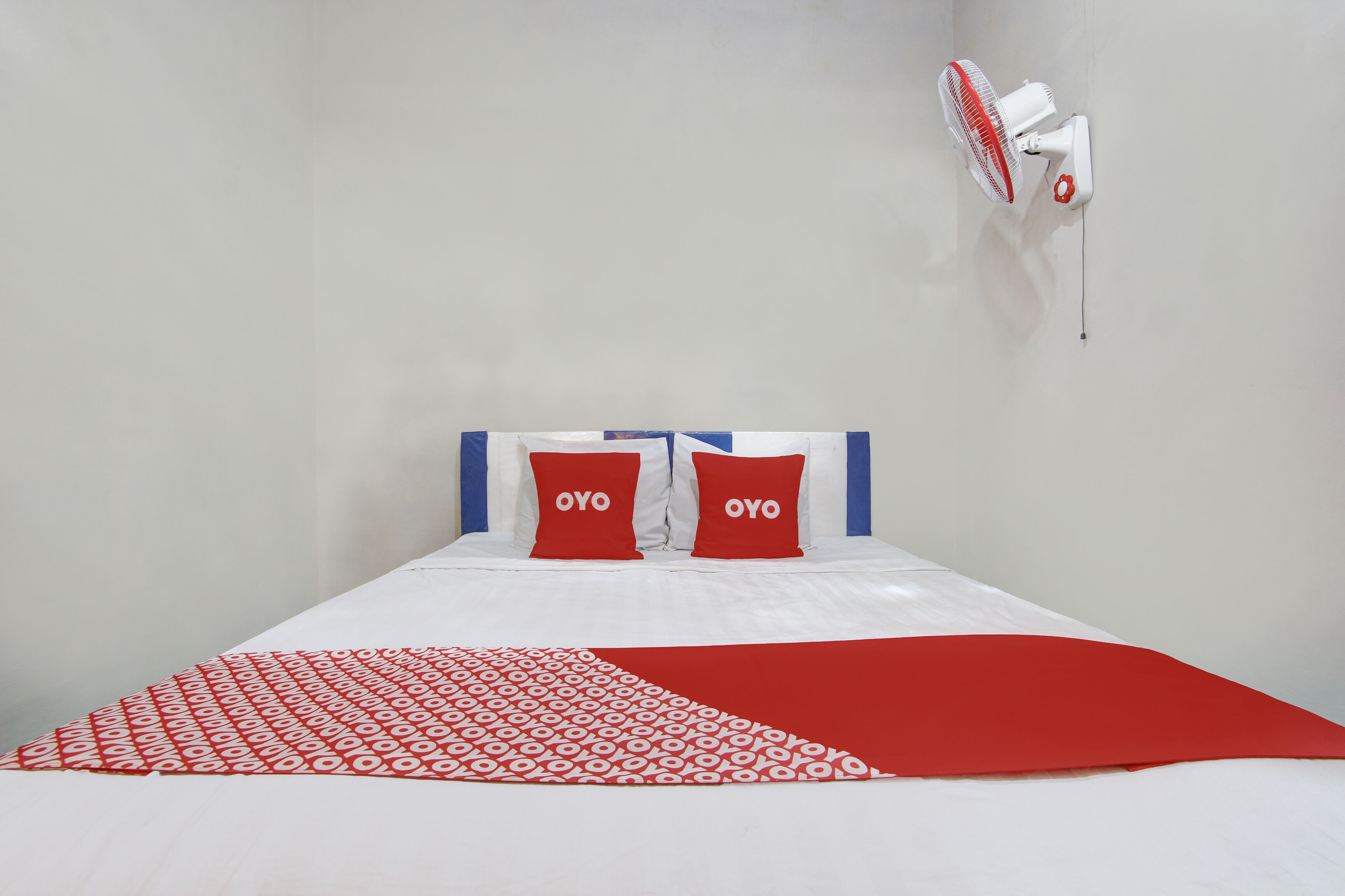 Uma cama com colcha estampada em branco e vermelho, duas almofadas vermelhas com a palavra "OYO" nelas e um ventilador fixado na parede.