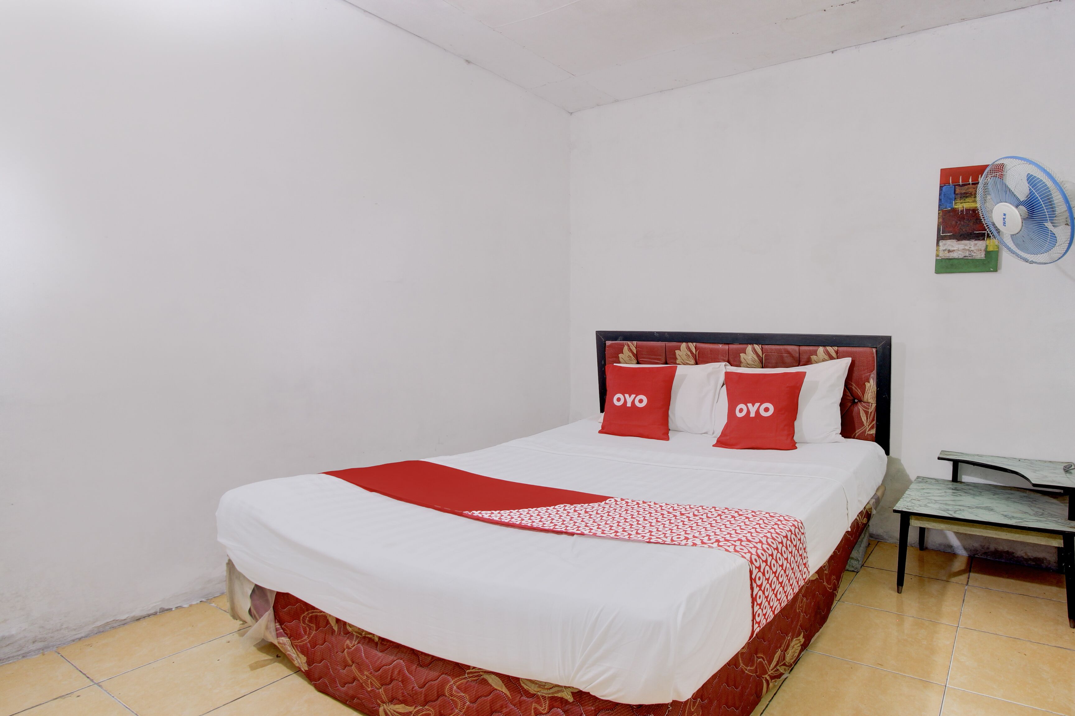 A neatly made bed with red and white bedding, a small table beside it, and a wall-mounted fan.