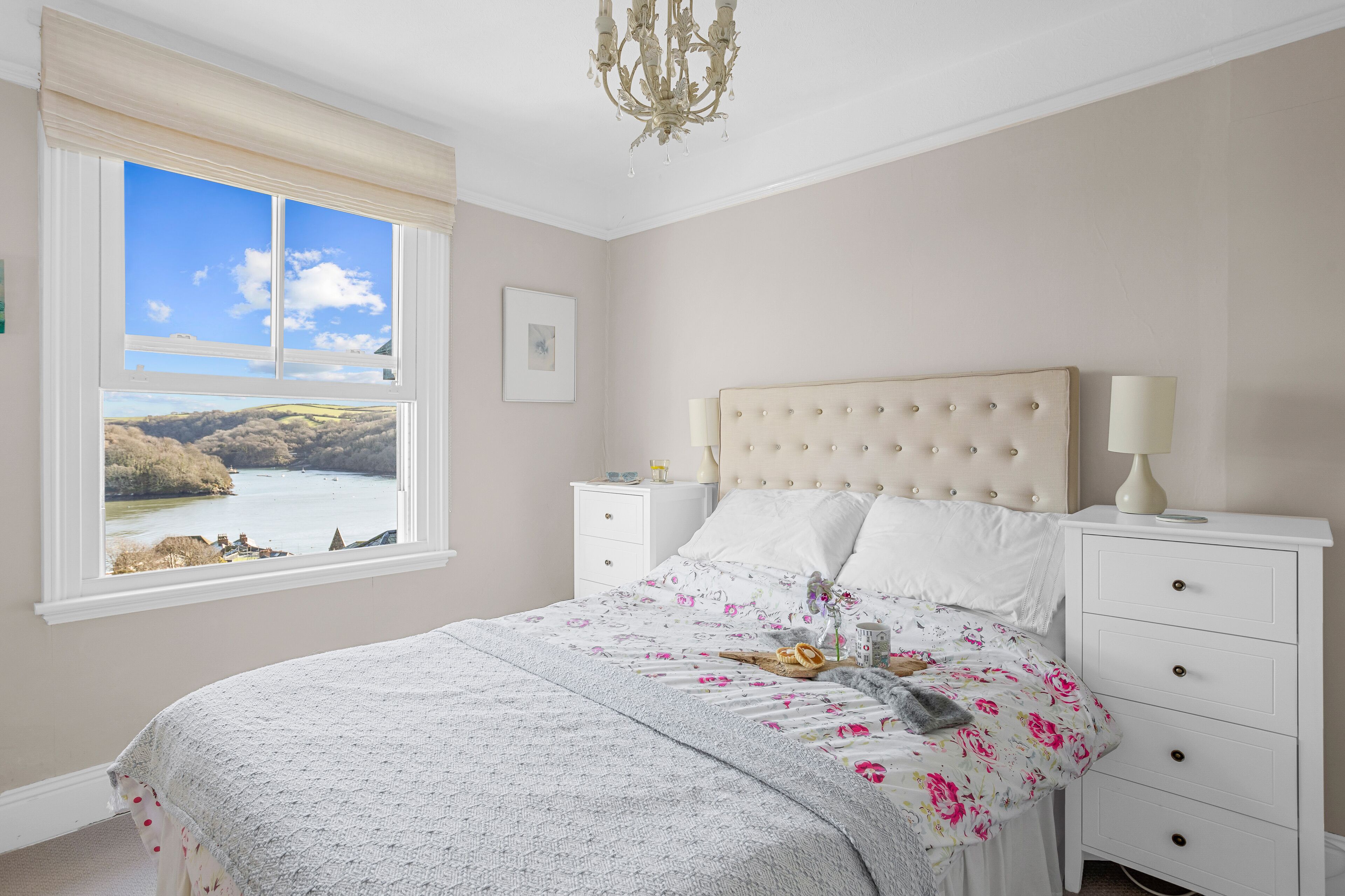 Une chambre avec une grande fenêtre, un lit recouvert d’une literie à motifs floraux, une tête de lit capitonnée et une table de chevet avec une lampe.