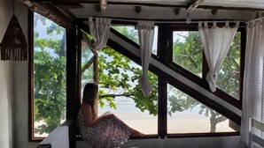 A room with a large window, a view of greenery, and a person sitting by the window. - Chalet Bromelias, Praia do Sono, Sea View (Paraty)