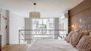 Une chambre moderne avec un grand lit, une vue sur la ville grâce à une grande fenêtre et un escalier menant à l’étage supérieur.