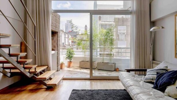 Un salon moderne avec un escalier, de grandes fenêtres et une vue sur des immeubles et des arbres.