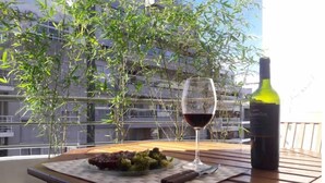 Une table à manger avec une assiette de nourriture, un verre à vin et une bouteille de vin, le tout placé sur un balcon d’où l’on peut admirer un bâtiment et des plantes de bambou.