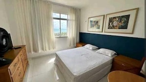 Habitación de hotel con cama, cómoda de madera, escritorio, silla, ventana con cortinas y dos cuadros en la pared.