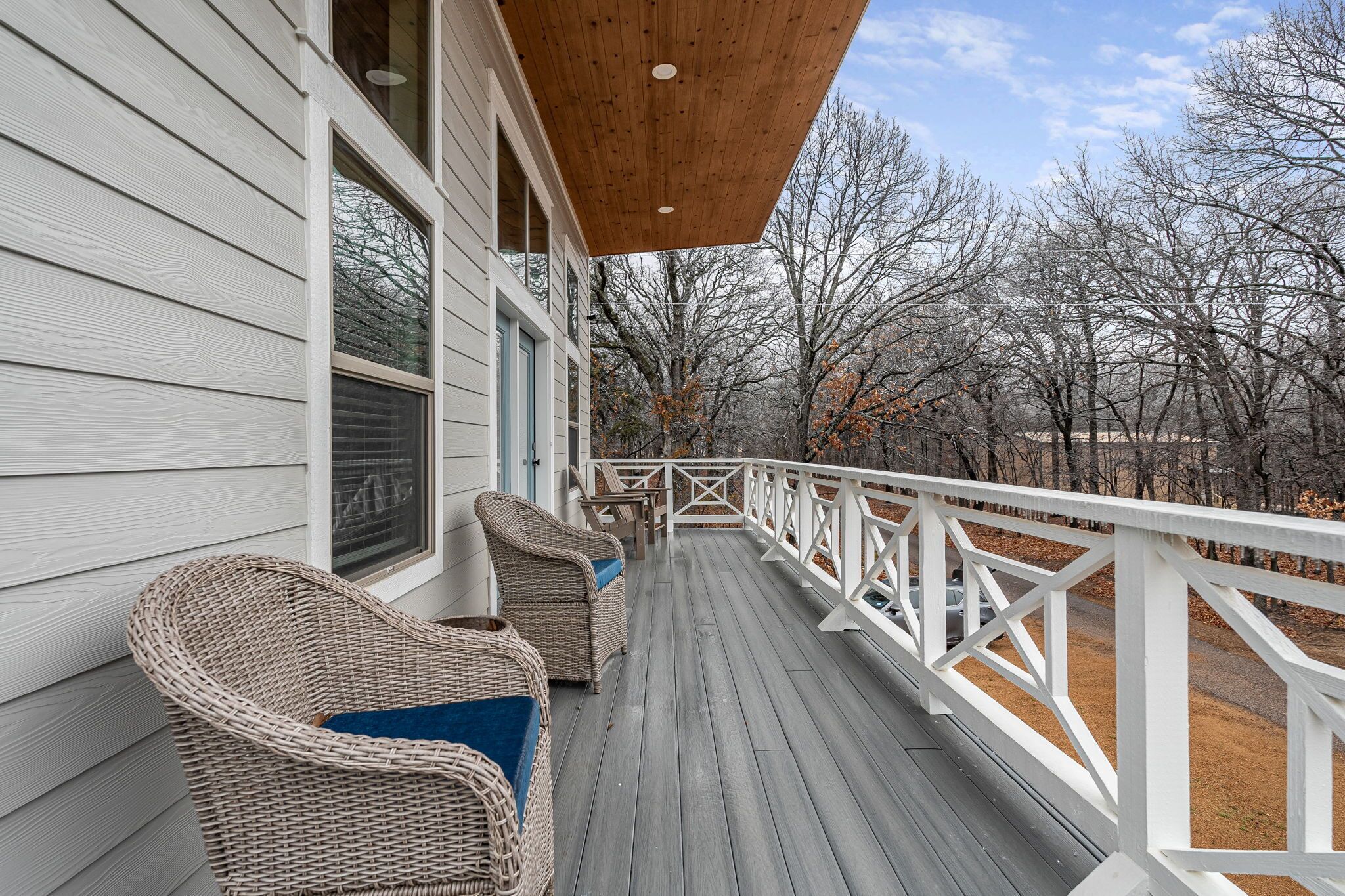 Une terrasse en bois avec des chaises en osier, une balustrade blanche et une vue sur les arbres.