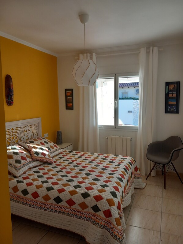 A bedroom with a bed, a chair, a window with curtains, and a radiator. - Villa Aucamar- Moraira (TEULADA MORAIRA)