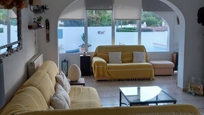 A living room with a yellow sofa, a glass coffee table, and a view through an arched doorway to the outside. - Villa Aucamar- Moraira (TEULADA MORAIRA)