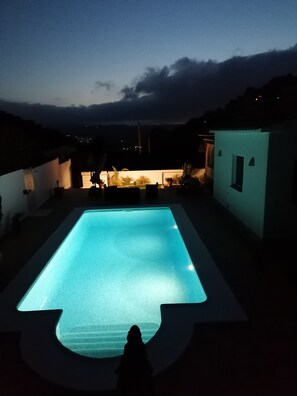 A swimming pool at dusk with surrounding buildings and a view of the landscape.